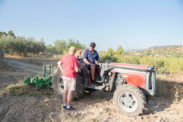 Dois fazendeiros mais velhos conversando no campo sobre um problema de trator, um deles sentado em um trator