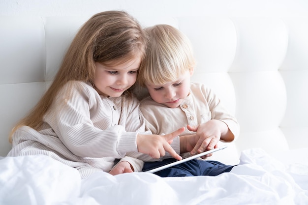 dois filhos bonitos menina e menino irmão e irmã usando tablet sentado na cama no quarto moderno claro