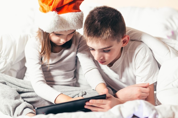 Dois filhos, um menino e uma menina, um irmão e uma irmã, amigos sentam-se debaixo de um cobertor branco na cama e brincam, lêem, estudam no tablet com um chapéu vermelho papai noel