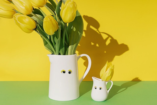 Dois frascos brancos com olhos de brinquedo grandes e pequenos com buquê de flores de tulipa amarela em um fundo amarelo e verde com sombras nítidas Composição floral criativa de primavera engraçada Papel de parede de Páscoa Decoração para casa