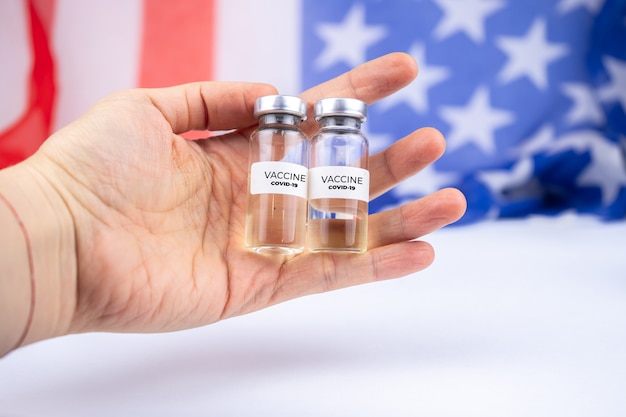 Dois frascos com a vacina covid-19 em uma das mãos. Bandeira dos Estados Unidos
