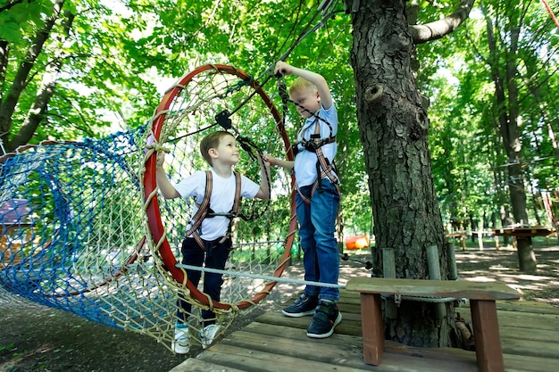 Foto dois garotinho com equipamento de escalada está caminhando ao longo de uma estrada de corda em um parque de aventura, segurando uma corda e um mosquetão.