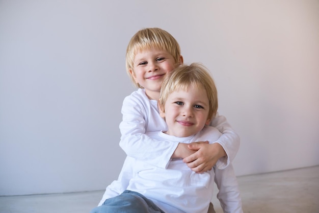 Dois garotos loiros sorrindo, gêmeos abraçados sentados no chão.