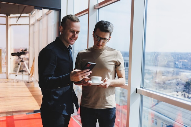 Dois gerentes homens de negócios ficam perto da janela em um escritório moderno e discutem um novo projeto dois homens com óculos estão falando sobre um novo negócio