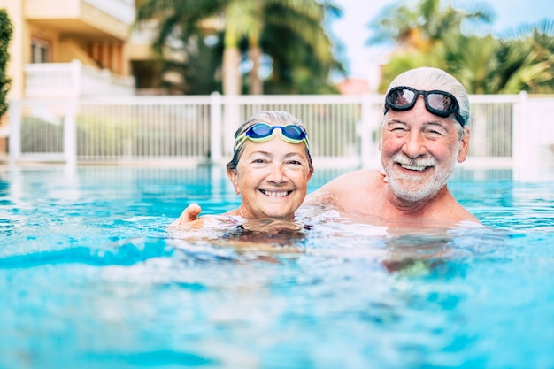 Dois giros idosos e aposentados na água da piscina se divertindo e curtindo juntos - duas pessoas maduras apaixonadas olhando para a câmera - fazendo exercícios e treinando juntos sorrindo