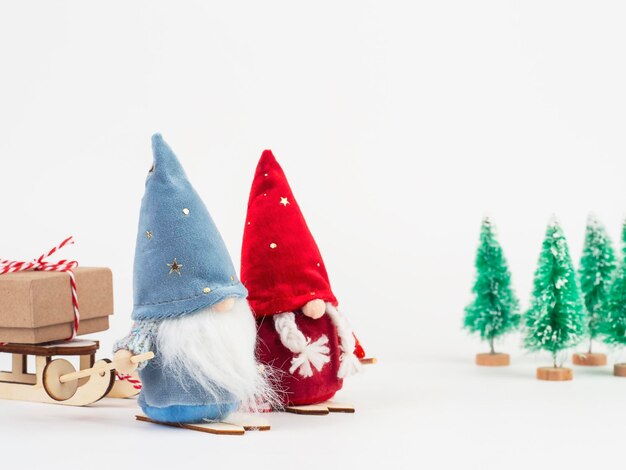 Dois gnomos escandinavos Nisse com esquis nas mãos estão carregando um trenó com um presente Há árvores de Natal ao fundo