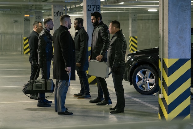 Dois grupos de assassinos de preto com revólveres e bolsas interagindo enquanto se posicionam frente a frente no estacionamento