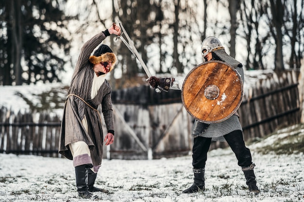 Dois guerreiros antigos em armadura com armas lutando com espadas na neve