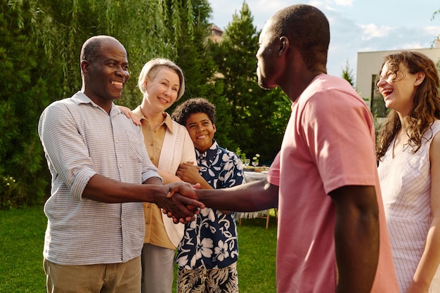 Dois homens afro-americanos felizes apertando as mãos enquanto estão na frente da câmera contra suas esposas e um lindo garoto e se cumprimentando