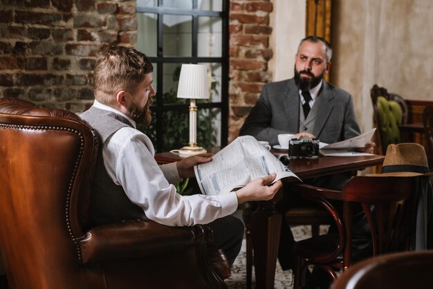 Dois homens barbudos vestindo ternos antiquados conversando ou discutindo algo no restaurante