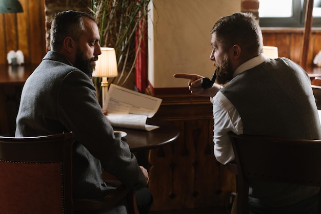 Dois homens barbudos vestindo ternos antiquados conversando ou discutindo algo no restaurante