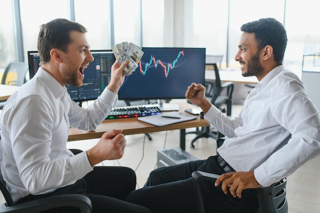 Dois homens comerciantes sentados à mesa no escritório juntos monitorando gráficos de vela de dados de ações na tela analisando o fluxo de preços sorrindo alegre tendo o conceito de trabalho em equipe de lucro