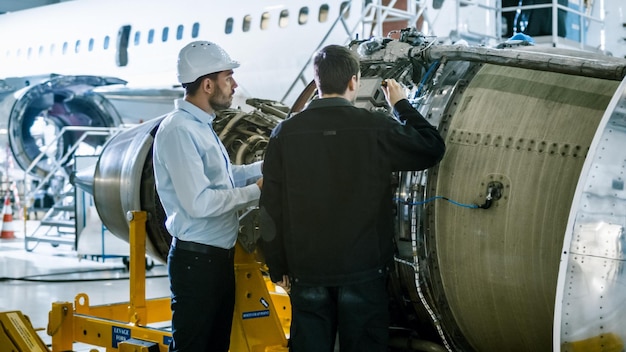 Dois homens de capacete estão em frente a um motor de avião.