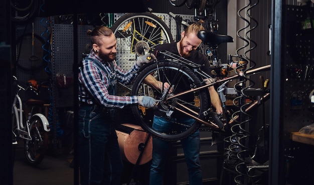 Dois homens elegantes sorridentes bonitos trabalhando com uma bicicleta em uma oficina. Os trabalhadores reparam e montam a bicicleta em uma oficina.
