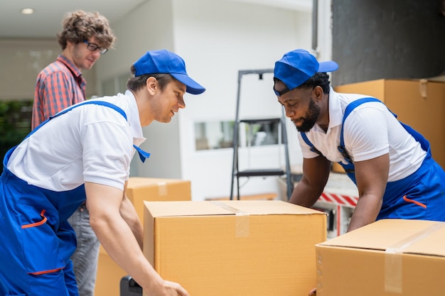 Dois homens em uniforme azul carregando caixa de papelão descarregando do caminhão de serviço de mudança