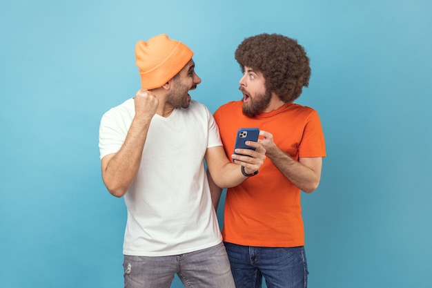 Dois homens espantados e animados em pé com o celular e comemorando o sucesso com os punhos cerrados