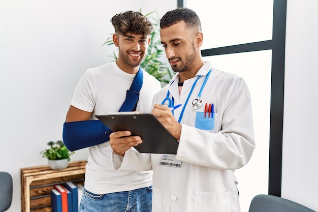 Foto dois homens hispânicos, médico e paciente escrevendo na prancheta em pé na sala de espera do hospital