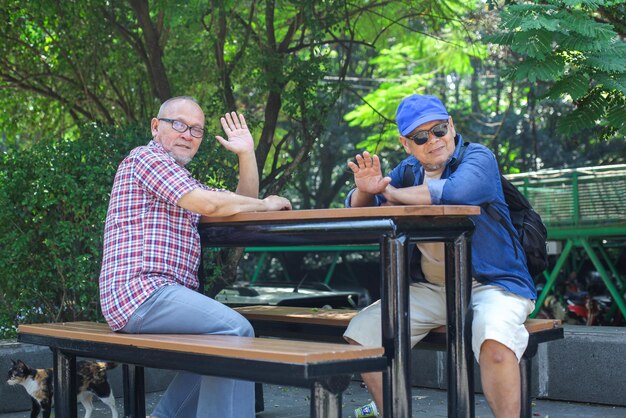Dois homens idosos amigáveis vestindo roupas casuais cumprimentando com as mãos acenando enquanto estão sentados ao ar livre