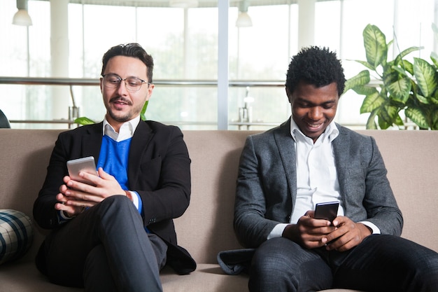 Dois homens multiculturais sentados no sofá segurando telefones celulares