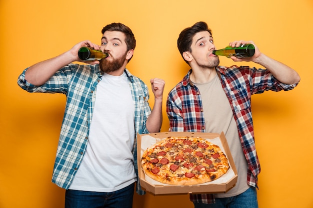 Dois homens satisfeitos em camisas, bebendo cerveja, segurando a parede amarela isolada de pizza