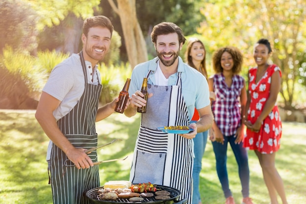 Dois homens segurando uma garrafa de cerveja enquanto prepara churrasqueira