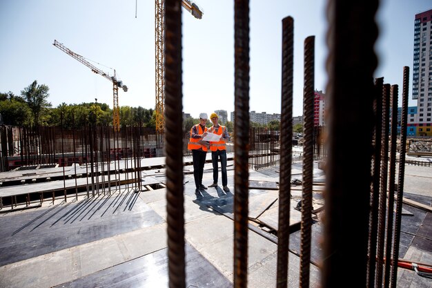 Dois homens vestidos com camisas, coletes de trabalho laranja e capacetes exploram a documentação da construção no canteiro de obras perto das estruturas de aço.