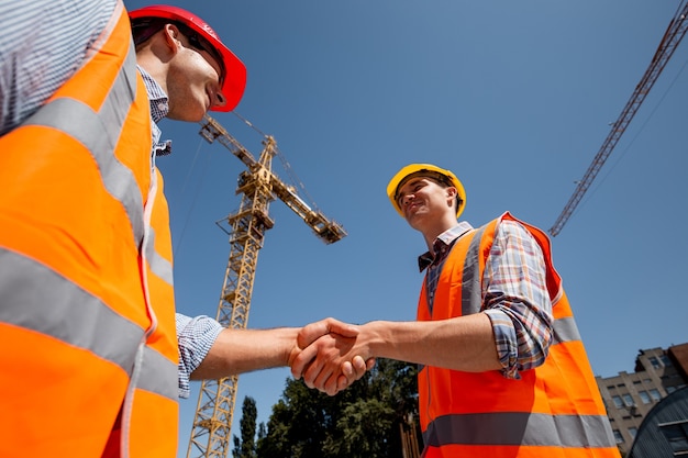 Dois homens vestidos com coletes de trabalho laranja e capacetes apertam as mãos no canteiro de obras perto do guindaste. .