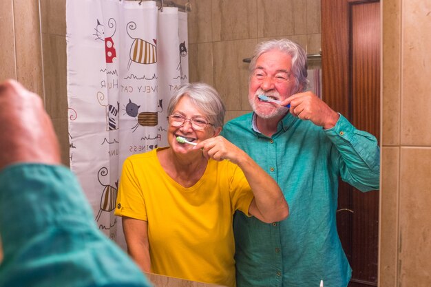 Dois idosos felizes e sorridentes, escovando os dentes um do outro juntos em casa no banheiro - cuidando de si e cuidando de si mesmos