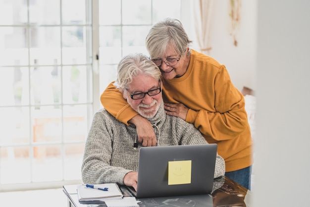 Dois idosos felizes trabalhando e usando laptop juntos em casa, sorrindo e se divertindo juntos - estilo de vida fechado