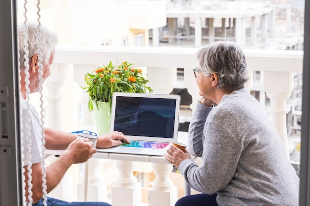 Dois idosos no terraço se divertindo e curtindo - homem com laptop mostrando algo para sua esposa - mulher com café e óculos olhando e sorrindo para o homem - ao ar livre