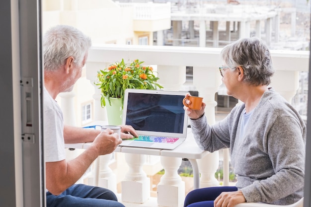 Dois idosos no terraço se divertindo e curtindo - homem com laptop mostrando algo para sua esposa - mulher com café e óculos olhando e sorrindo para o homem - ao ar livre