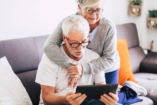 Dois idosos sorrindo e olhando para o mesmo tablet abraçados no sofá - conceito interno, em casa - caucasianos maduros e aposentados e mulheres usando tecnologia