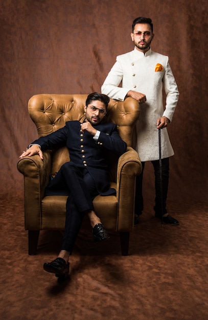 Dois indianos usam roupas étnicas ou tradicionais, modelos masculinos com sherwani ou pijama kurta, sentados e posando em uma poltrona ou sofá contra um fundo marrom grunge, foco seletivo