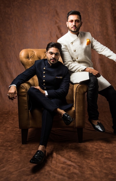 Dois indianos usam roupas étnicas ou tradicionais, modelos masculinos com sherwani ou pijama Kurta, sentados e posando em uma poltrona ou sofá contra um fundo marrom grunge, foco seletivo