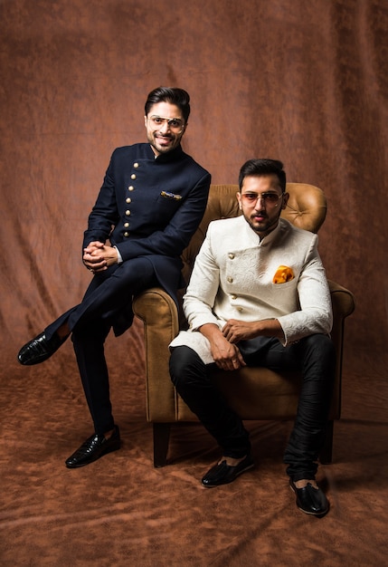 Dois indianos usam roupas étnicas ou tradicionais, modelos masculinos com sherwani ou pijama Kurta, sentados e posando em uma poltrona ou sofá contra um fundo marrom grunge, foco seletivo