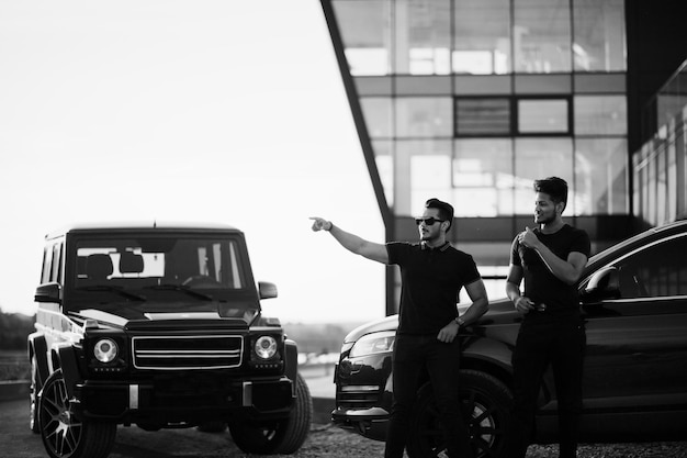 Dois irmãos asiáticos vestem todo preto posando perto de carros suv.