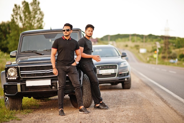 Dois irmãos asiáticos vestem todo preto posando perto de carros suv.