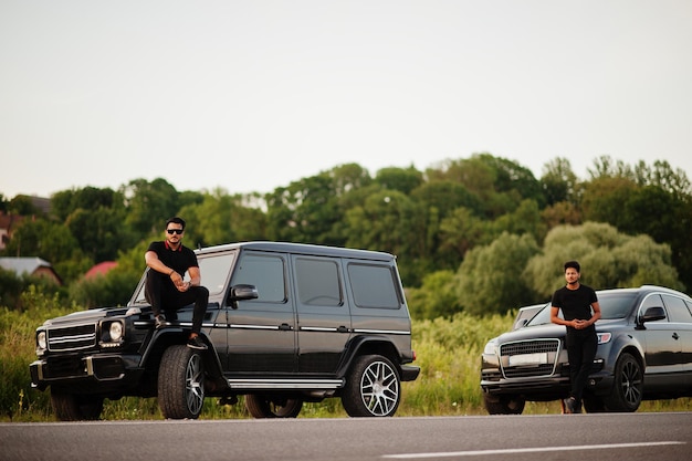 Dois irmãos asiáticos vestem todo preto posando perto de carros suv.