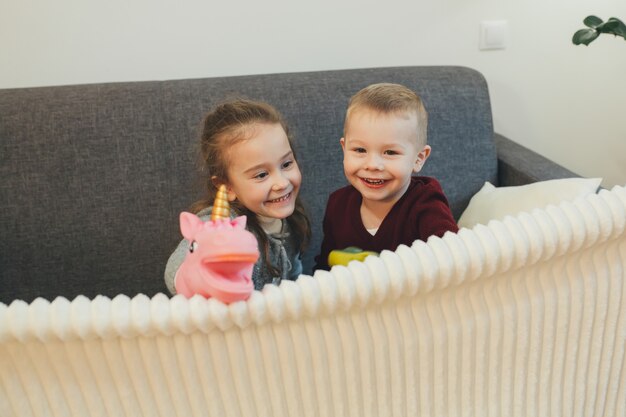 Dois irmãos caucasianos brincando no sofá com um unicórnio e um sorriso doce para a câmera