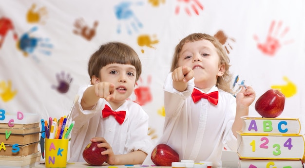 Dois irmãos de 7 e 4 anos vestidos com camisas brancas com laços vermelhos estão aparecendo em algo com o dedo.