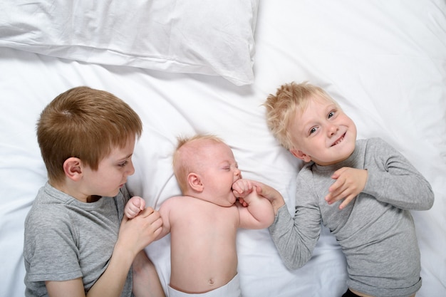 Dois irmãos mais velhos brincam e riem com o bebê mais novo em uma cama branca.