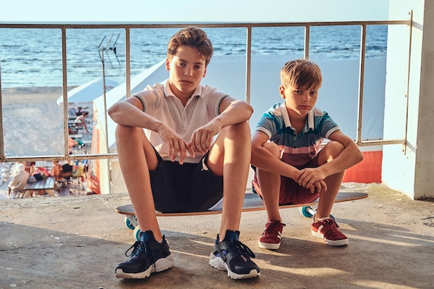 Dois irmãos sentados juntos em um skate perto do guardrail no contexto de um litoral.