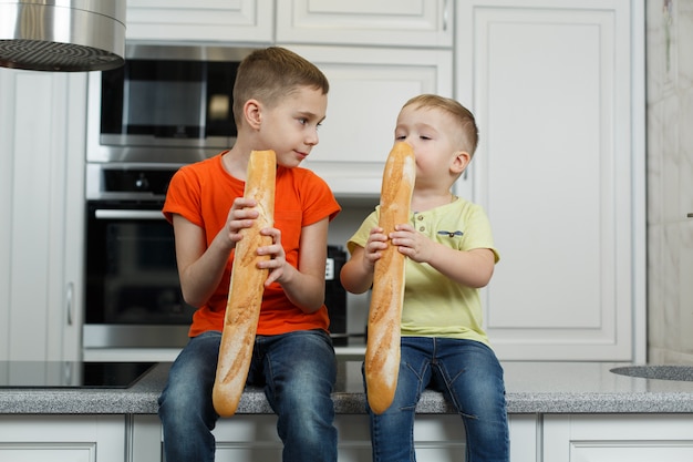 Dois irmãos tomam café da manhã na cozinha. garotos engraçados comendo um pão na cozinha. bebê fofo comendo baguete.