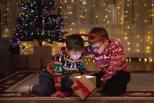 Dois irmãozinhos caucasianos abrindo os presentes de natal em casa perto da árvore de natal. irmãos felizes sentados no chão, olhando para uma caixa de presente no natal.