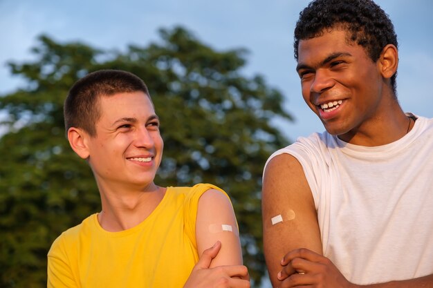 Dois jovens amigos afro-americanos e caucasianos sorridentes, mostrando as mãos com emplastros após a vacinação ao pôr do sol no verão