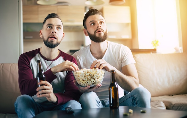 Dois jovens amigos barbudos felizes assistindo TV ou alguma partida de esporte enquanto estão sentados no sofá em casa no fim de semana, bebendo cerveja e comendo lanches