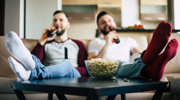 Dois jovens amigos barbudos felizes assistindo TV ou alguma partida de esporte enquanto estão sentados no sofá em casa no fim de semana, bebendo cerveja e comendo lanches