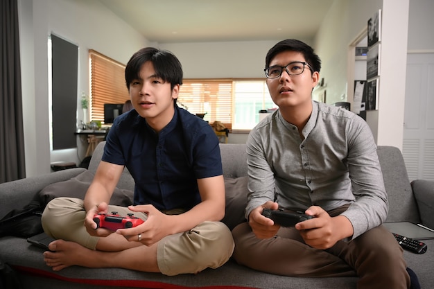 Dois jovens asiáticos jogando videogame enquanto está sentado no sofá em casa.