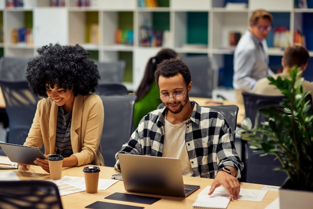 Dois jovens colegas de negócios diversos usando tecnologias modernas enquanto estão sentados à mesa e