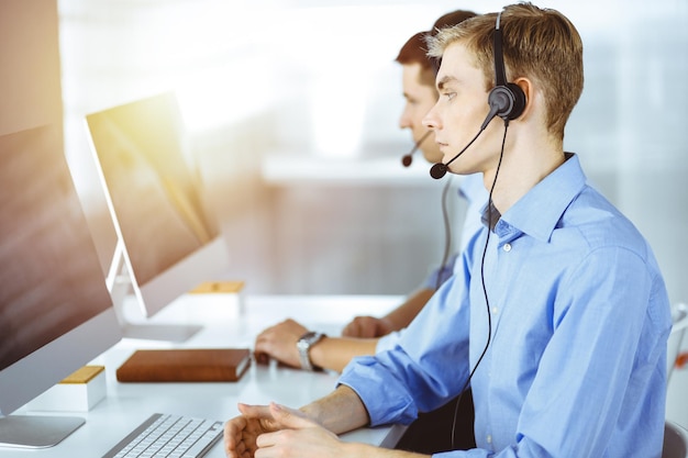 Dois jovens com fone de ouvido, sentados à mesa em um escritório moderno e ensolarado, ouvindo os clientes. Operadores de call center no trabalho.